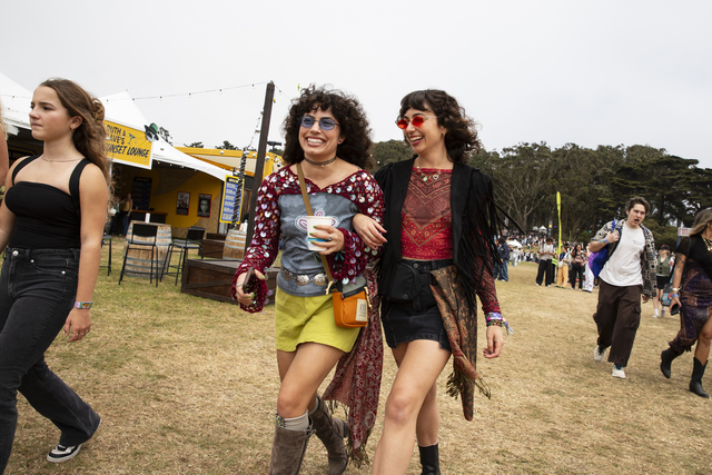 Two women wearing colorful, bohemian outfits and sunglasses walk arm in arm, smiling and laughing at an outdoor festival with other people nearby.